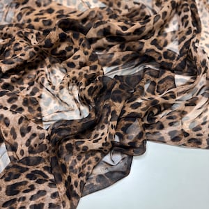 Könnte beinhalten: Ein fließender Stoff mit Leopardenmuster. Das Material ist ein leichter, transparenter Chiffon in Braun- und Schwarztönen. Der Stoff ist drapiert und gefaltet, wodurch ein Gefühl von Bewegung und Textur entsteht.