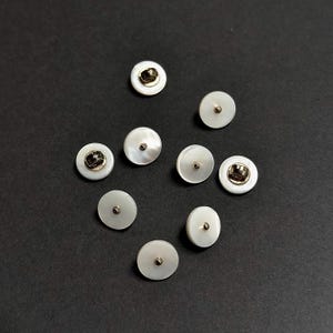 May include: Nine white round buttons with a gold centre. The buttons are arranged in a circle on a black surface.