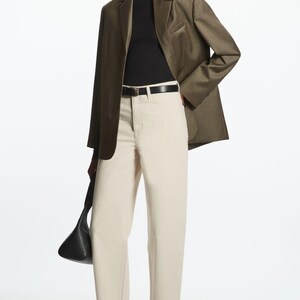 May include: An olive green blazer worn over a black turtleneck, paired with off-white high-waisted trousers, a black leather belt, black loafers, and a black handbag. The ensemble features structured tailoring and neutral tones.