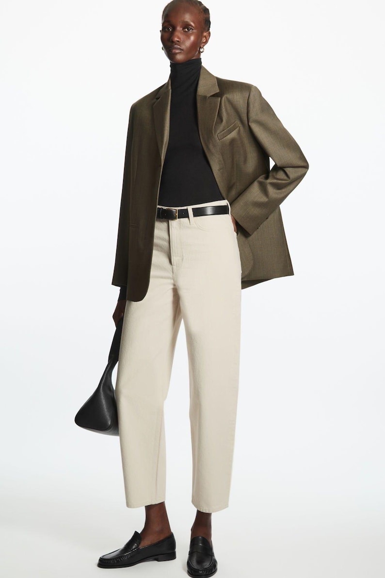 May include: An olive green blazer worn over a black turtleneck, paired with off-white high-waisted trousers, a black leather belt, black loafers, and a black handbag. The ensemble features structured tailoring and neutral tones.