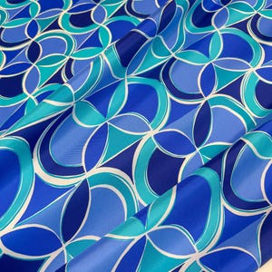 May include: A close-up of a fabric with a repeating geometric pattern in shades of blue and white. The design features curved shapes and lines, creating a flowing, abstract design. The fabric has a glossy sheen, suggesting a silk or satin material.