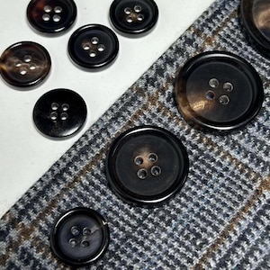 以下が含まれることがあります： 4つの穴が空いた茶色と黒の角ボタンが8つ集まったセット。ボタンは模様のある布の上に半円形に並べられています。