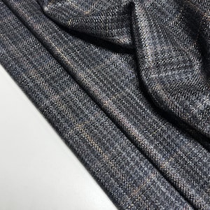 Op de afbeelding: Close-up van een gevouwen stof met een donkergrijs en blauw ruitpatroon. De stof heeft een getextureerde uitstraling met subtiele hints van bruine en witte draden die erdoorheen zijn geweven. Het materiaal lijkt een zwaar gewicht te hebben, geschikt voor kleding.