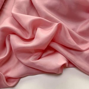 Puede incluir: Una pieza de tela rosa claro con una textura suave y satinada. El material está drapeado en pliegues suaves y fluidos, creando sombras y reflejos profundos en la superficie que enfatizan la calidad lustrosa y reflectante del tejido.