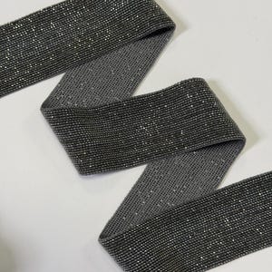 Può includere: Un nastro elastico grigio scuro con una superficie scintillante e brillante. Il nastro è piegato e curvo, mostrando la sua flessibilità. Il materiale sembra essere densamente ricoperto di piccoli elementi riflettenti, creando un effetto cangiante. Ideale per l'artigianato e gli abbellimenti.