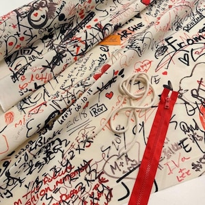 May include: White fabric with black and red graffiti-style writing and hearts. A red zipper and a white cord are visible in the bottom right corner.
