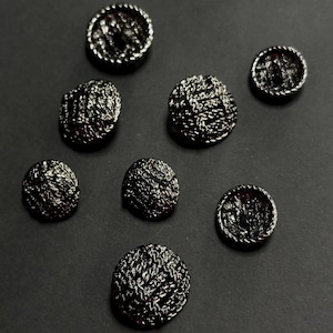 May include: Seven circular buttons with a dark metallic finish and a textured, woven-like surface pattern. Each button features a raised, rope-style border around the edge, creating a consistent appearance across the entire set of fasteners.