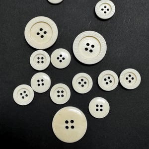 Puede incluir: Una colección de 12 botones redondos de plástico blanco con cuatro agujeros cada uno. Los botones están dispuestos en semicírculo sobre un fondo negro.