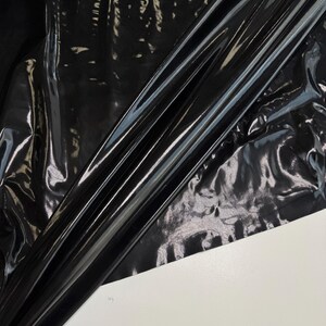 Peut inclure: Gros plan sur une feuille de tissu vinyle noir et brillant. Le matériau est plié et drapé sur une surface blanche, créant des reflets et des ombres. Le tissu a une finition brillante et réfléchissante.