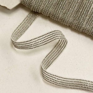 Könnte beinhalten: Silberne Perlenborte mit einem durchsichtigen, netzartigen Rücken. Die Borde besteht aus kleinen, runden Perlen, die gleichmäßig verteilt und in Reihen angeordnet sind.