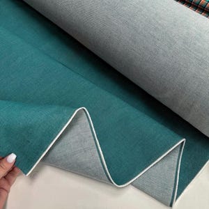 Puede incluir: Un rollo de tela verde azulado con un borde blanco, que muestra la textura y el color del material. La tela está doblada, revelando una parte inferior gris más clara. Una segunda tela con un estampado de cuadros es visible en el fondo.