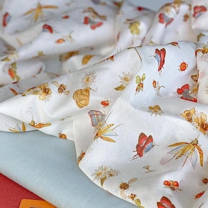 Pode incluir: Tecido branco com um padrão repetido de insetos e flores coloridas. O design inclui borboletas, abelhas, joaninhas e margaridas em tons de vermelho, amarelo e azul. O tecido é drapeado sobre uma superfície azul claro e vermelha.