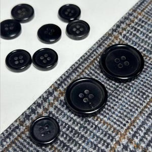May include: A close-up of ten black buttons with four holes each. The buttons are arranged in a semi-circle on a white surface. The buttons are made of a smooth, shiny material.