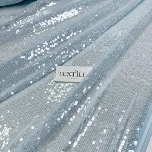 May include: Light blue sequin fabric with a shimmering effect. The fabric is draped, showing folds and texture. A white label with the text "TEXTILE LIBRARY" is visible.