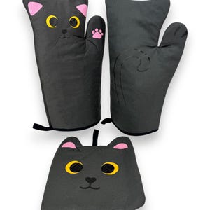 May include: Two black oven mitts and one matching pot holder shaped like cat heads. The mitts feature yellow eyes, pink inner ears, and a pink paw print on the thumb. The pot holder displays yellow eyes and pink inner ears with a black hanging loop.