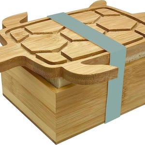 May include: A bamboo box with a turtle-shaped lid. The lid is light brown with a carved turtle design. A light blue band wraps around the box. The box is made of bamboo and has a square shape.
