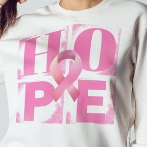 Puede incluir: Sudadera blanca con una cinta rosa y la palabra "HOPE" impresa en letras rosas.