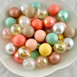 May include: A collection of colorful round beads in a white bowl. The beads are in various colors, including pink, peach, green, yellow, and iridescent. Some beads have a glittery texture, while others are smooth and solid. The beads are likely for jewelry making or crafts.