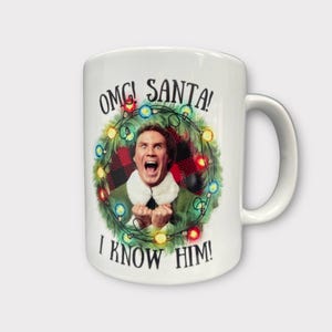 May include: White ceramic mug with a Christmas-themed design featuring Buddy the Elf from the film Elf. The mug has a red and black plaid background with a green wreath of Christmas lights and the text "OMG! SANTA! I KNOW HIM!"
