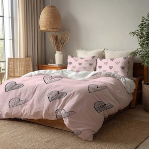Può includere: Un copripiumino rosa e due federe con un motivo a cuore nero. La biancheria da letto è su una struttura letto in legno. La stanza ha una palette di colori neutri con una lampada a sospensione intrecciata e una sedia intrecciata.