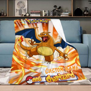 May include: A soft blanket with a vibrant design featuring the Pokemon Charizard. The blanket is predominantly orange and yellow, with a blue-winged Charizard illustration and the Pokemon logo. Includes a custom name section.