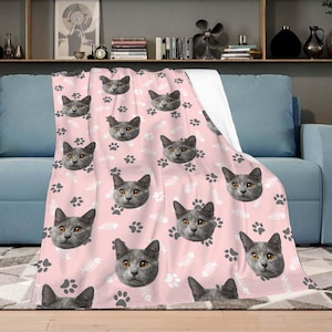 Op de afbeelding: Roze fleece deken met een herhalend patroon van een grijs kattengezicht, witte poot afdrukken en witte visgraten.