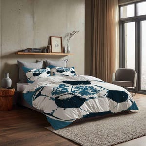 Op de afbeelding: Een wit en blauwgroen voetbalthema beddengoed set. De dekbedovertrek en kussenslopen hebben een groot voetbalontwerp. Het bed is opgemaakt met een grijs hoofdbord en een neutraal gekleurd tapijt.