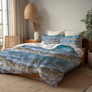 May include: A bed dressed with a duvet cover and two pillowcases, all with a realistic beach scene print. The design features clear blue water, white foam, and pebbles. The bed is wooden and rests on a woven rug.