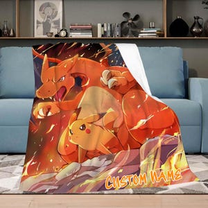 Puede incluir: Una manta suave con una vibrante ilustración de Charizard y Pikachu en tonos naranja, rojo y amarillo. La manta tiene el texto "CUSTOM NAME" en una fuente estilizada. La manta está sobre un sofá azul.