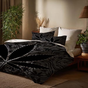 Könnte beinhalten: Schwarzes und graues Bettwäsche-Set mit einem großen Cannabisblatt-Design. Das Set beinhaltet einen Bettbezug und zwei Kissenbezüge. Das Design hat ein detailliertes, strukturiertes Aussehen. Das Bett ist mit weißen Laken und Kissen bezogen.
