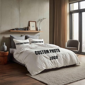 Könnte beinhalten: Ein weißes Bettwäsche-Set mit dem Text "CUSTOM PHOTO LOGO" auf dem Bettbezug und den Kissenbezügen. Die Bettwäsche befindet sich auf einem Bett mit grauem Rahmen, vor einer neutralfarbenen Wand und einem Fenster mit transparenten Vorhängen.