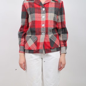 May include: A long-sleeve button-up jacket featuring a red, charcoal grey, and beige plaid pattern. The jacket includes a pointed collar, four front buttons, and two square patch pockets at the waist. The sleeves have buttoned cuffs.