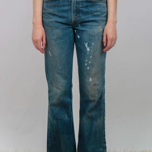 May include: High-waisted blue denim jeans featuring a faded wash, a classic five-pocket design, copper-toned rivets, and visible white paint splatter marks on the front of the legs, with a straight-leg cut and a button fly closure.