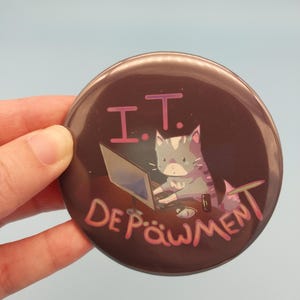 May include: A circular, brown button with the pink text "I.T. Depäwment". The button features a cartoon cat working on a laptop. The cat is grey and white with pink accents.