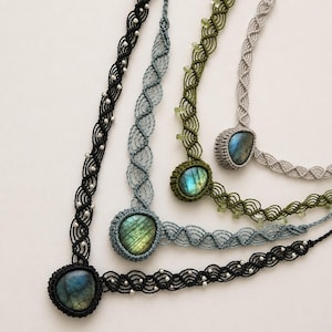 Può includere: Quattro collane in macramè con pietre di labradorite ovali incastonate in cordini annodati. Le collane sono nere, azzurre, verde oliva e grigio chiaro, ognuna decorata con piccole perline integrate negli intricati motivi di tessitura.