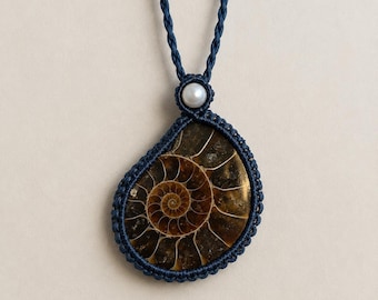 May include: A pendant featuring a polished brown ammonite fossil encased in dark blue macrame cord. The fossil displays a spiral chambered pattern. A single white pearl is positioned at the top of the pendant, connecting to a braided cord necklace.