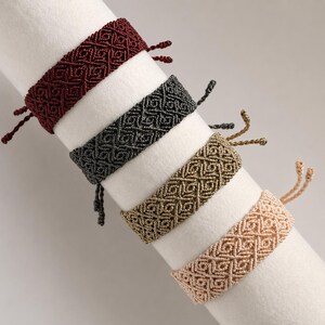 Könnte beinhalten: Vier gewebte Armbänder mit einem sich wiederholenden geometrischen Rautenmuster. Die Armbänder sind in Dunkelrot, Anthrazit, Metallic-Gold und Hellbeige gehalten. Jedes Armband verfügt über verstellbare Enden mit Knoten.