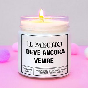 Può includere: Una candela bianca accesa in un barattolo di vetro trasparente con un'etichetta in bianco e nero. L'etichetta recita "IL MEGLIO DEVE ANCORA VENIRE" e "SPICY WAX CANDLE". La candela è su una superficie rosa con pompon rosa e bianchi.