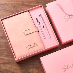 May include: A pink gift set containing a notebook with a gold-toned rectangular clasp and the name Stella embossed on the cover, a matching pink pen with the name Stella, and a pen refill, all housed in a pink box with a matching gift bag.