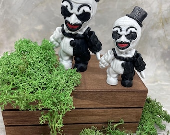 May include: Two small, white and black clown figurines with top hats and red smiles. They stand on a wooden box, surrounded by green moss. The figurines have a classic clown design.