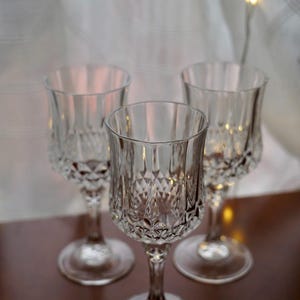 May include: Three clear crystal wine glasses with diamond-cut detailing. The glasses have a classic design with a stemmed base and a wide bowl. The glasses are arranged on a dark brown surface, with a soft, white background.