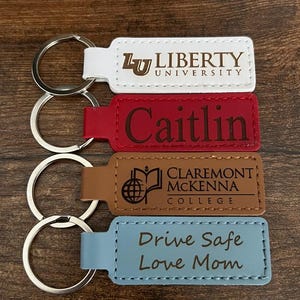 May include: Four keychains with silver key rings. The keychains are rectangular with rounded corners and are white, red, brown, and light blue. Text on the keychains includes "Liberty University", "Caitlin", "Claremont McKenna College", and "Drive Safe Love Mom".