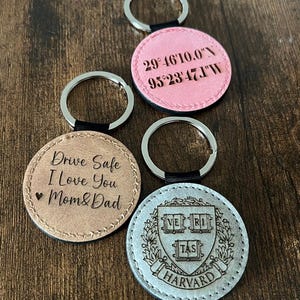 Puede incluir: Una colecci&oacute;n de llaveros redondos con aros plateados. Un llavero es de color marr&oacute;n claro con el texto "Drive Safe I Love You Mom & Dad". Otro es rosa con coordenadas. El &uacute;ltimo es plateado con un escudo de Harvard.