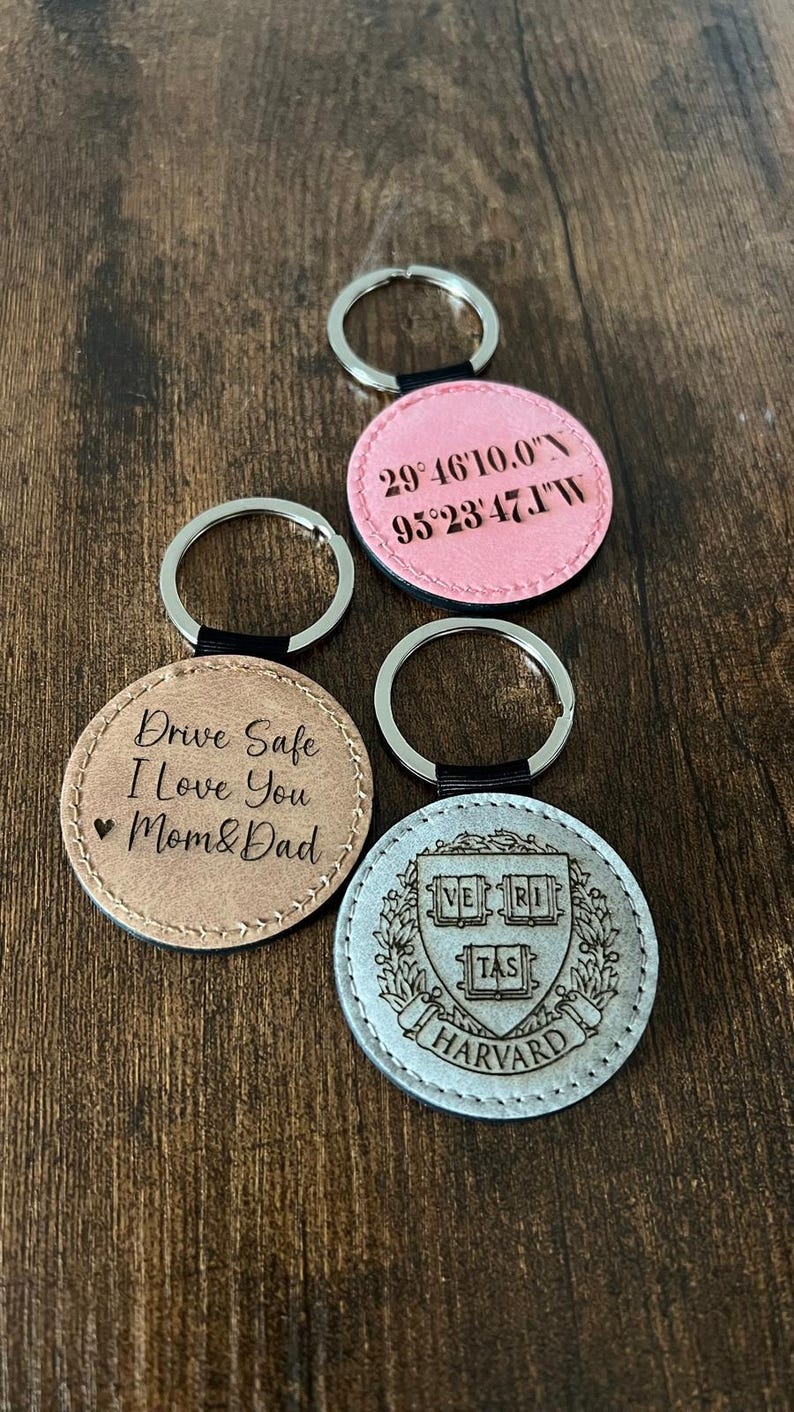 Puede incluir: Una colecci&oacute;n de llaveros redondos con aros plateados. Un llavero es de color marr&oacute;n claro con el texto "Drive Safe I Love You Mom & Dad". Otro es rosa con coordenadas. El &uacute;ltimo es plateado con un escudo de Harvard.