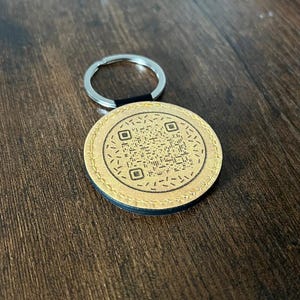 Puede incluir: Un llavero redondo de cuero color canela con un borde negro y un anillo plateado. El llavero presenta un dise&ntilde;o de c&oacute;digo QR en el centro. El fondo es una superficie de madera marr&oacute;n oscuro.