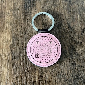 Puede incluir: Un llavero redondo rosa con un c&oacute;digo QR en el centro. El llavero tiene un llavero plateado y una correa negra que conecta el anillo con el c&iacute;rculo rosa. El fondo es una superficie de madera.
