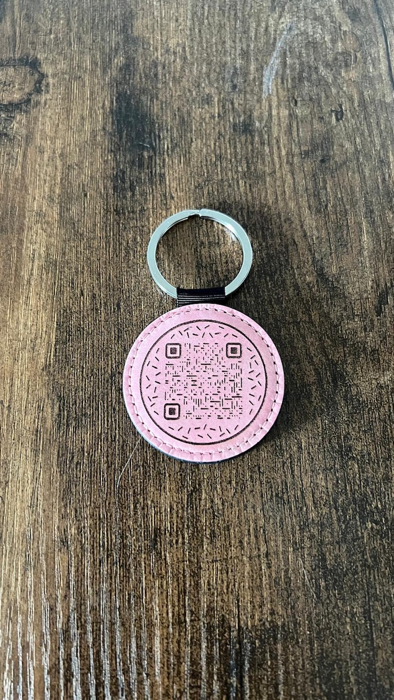 Puede incluir: Un llavero redondo rosa con un c&oacute;digo QR en el centro. El llavero tiene un llavero plateado y una correa negra que conecta el anillo con el c&iacute;rculo rosa. El fondo es una superficie de madera.