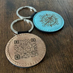 Puede incluir: Dos llaveros con colgantes redondos de cuero. Uno es marr&oacute;n claro con un c&oacute;digo QR, el otro es turquesa con un dise&ntilde;o de br&uacute;jula. Ambos tienen llaveros plateados y detalles en negro. Sobre una superficie de madera.