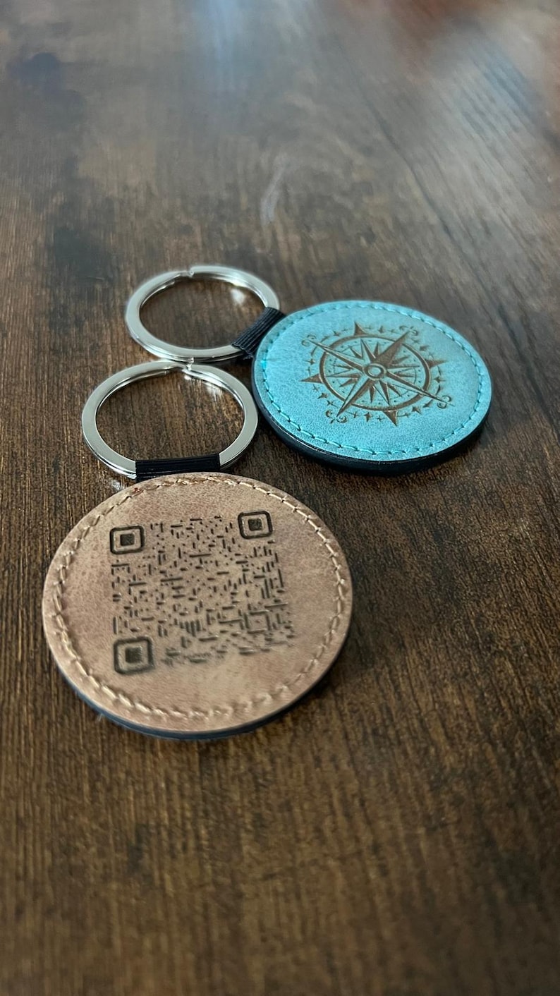 Puede incluir: Dos llaveros con colgantes redondos de cuero. Uno es marr&oacute;n claro con un c&oacute;digo QR, el otro es turquesa con un dise&ntilde;o de br&uacute;jula. Ambos tienen llaveros plateados y detalles en negro. Sobre una superficie de madera.