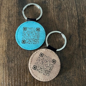 Puede incluir: Dos llaveros redondos con c&oacute;digos QR. Uno es azul claro, el otro es beige. Ambos tienen llaveros plateados y detalles en cuero negro. Los c&oacute;digos QR son negros e impresos en la parte delantera. Sobre una superficie de madera oscura.